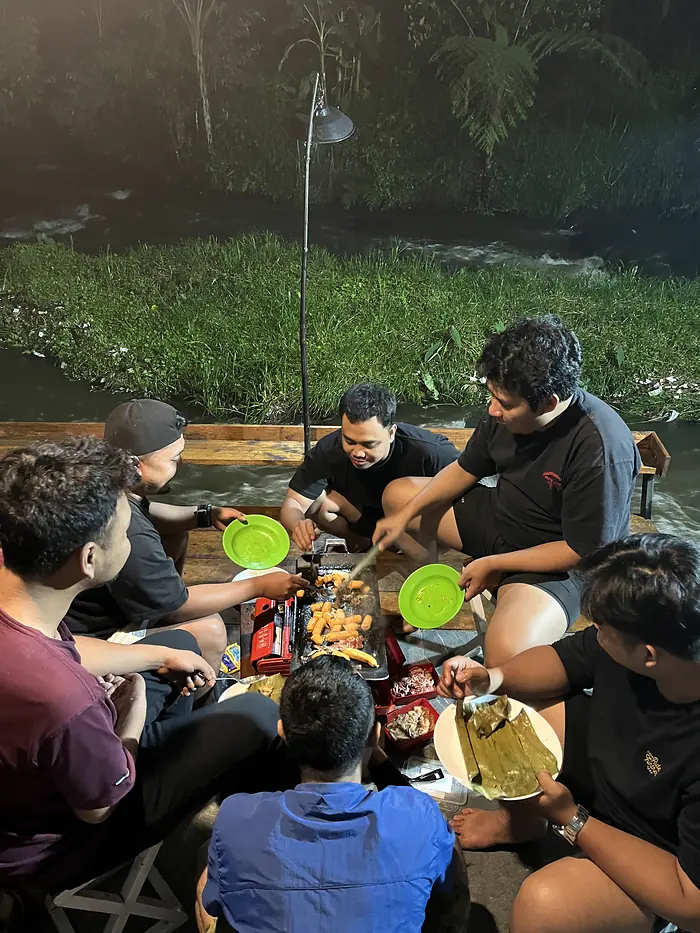 Tamu makan bersama di meja panjang tepi sungai pada malam hari