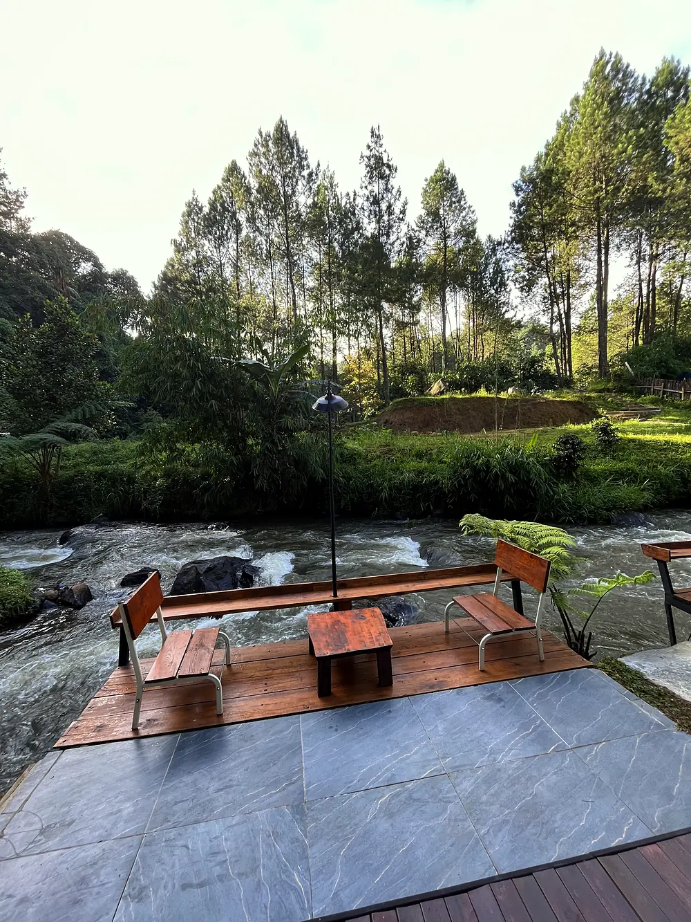 Deck kayu dengan kursi menghadap sungai berarus, dikelilingi hutan pinus Pangalengan
